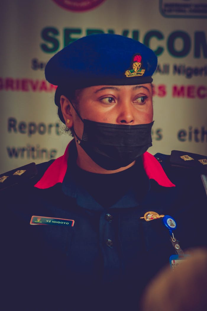 A female security officer stands in uniform with a focused expression, wearing a mask.